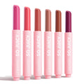 Six ColourPop So Juicy Plumping Gloss Balm with Peptides sticks, So Balm, stand upright and uncapped on a white background, displaying fuchsia, coral, rosy nude, beige, warm brown, and deep berry in pink tubes labeled SO JUICY.