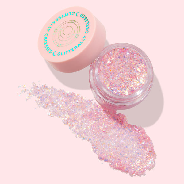 Moonlight Legend ColourPop Glitter Gel spreads from an open clear jar, showcasing iridescent pink and opal chunky glitter flakes; pale pink lid with celestial graphic sits beside on a soft pink backdrop, ideal for face and body sparkle.