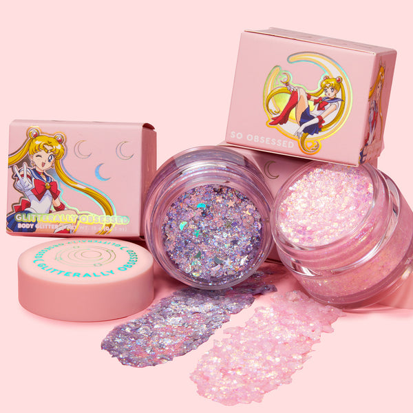 Moon Prism Power ColourPop Glitter Gel jars, lids off, show lilac and pink iridescent chunky glitter swatched on a pastel pink surface, surrounded by Sailor Moon–themed pink boxes, ideal for face and body sparkle.