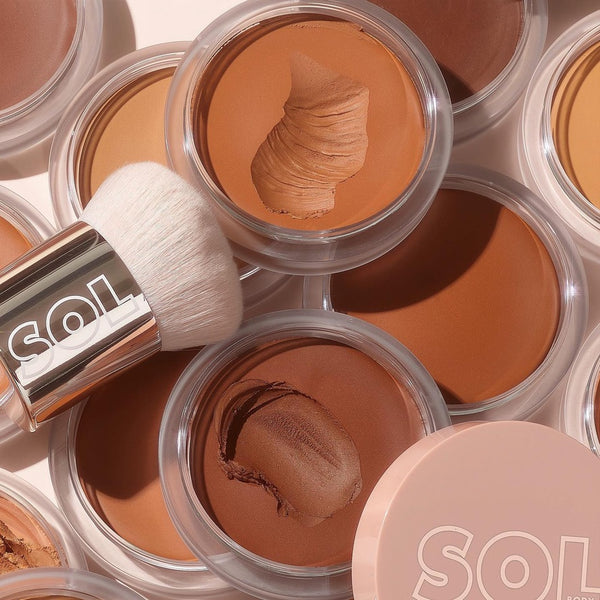 ColourPop SOL Face & Body Bronzing Balm in Light swatched in a clear round pot, showing its creamy tan texture, arranged with other shade pots and a SOL-branded buffing brush on a soft beige backdrop for sun-kissed face and body.