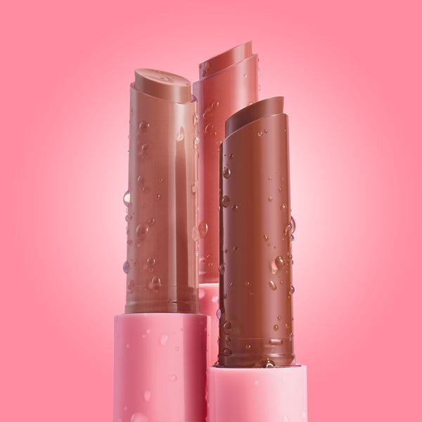 Three Lush Pout bullet balms stand upright with water droplets, displaying nude and rosy tints in pink tubes on a soft pink gradient - ColourPop So Juicy Plumping Gloss Balm with Peptides for a glossy, plumped finish.