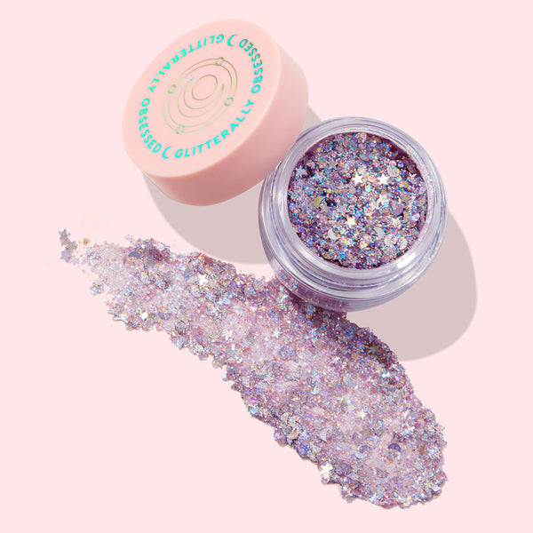 Open jar of Moon Prism Power ColourPop Glitter Gel showing chunky iridescent lavender flakes and star confetti, with a wide swatch spread beside it; peach lid with teal “Glitterally Obsessed” logo on a pale pink background.