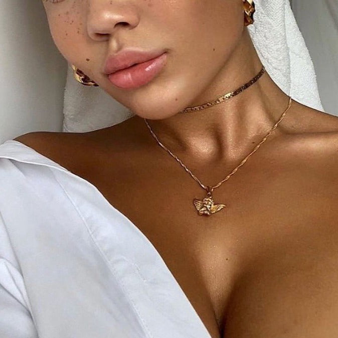 Bronzed collarbones and shoulders glow with the Golden shade of ColourPop SOL Glow Oil, showcasing a dewy, luminous finish as a model wears a white off-shoulder shirt, gold hoop earrings, a choker, and a bee pendant in soft indoor light.