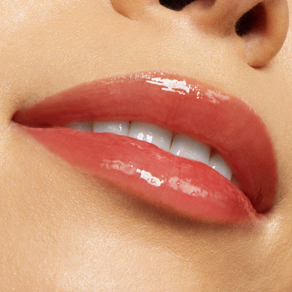 Glossy, coral-pink lips wearing Vampy Lips Lux Lip Oil, showcasing a sheer, high-shine finish from ColourPop Lux Lip Oil as the mouth softly smiles with teeth peeking, close-up on smooth, hydrated skin.