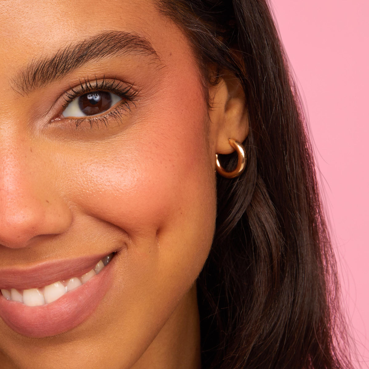 Smiling modelβs left cheek and lips wearing Mood 24/7 ColourPop Cheek & Lip Set, showing dewy peach blush and rosyβnude lip tint with a soft glow; a gold hoop earring and dark hair frame the face against a pink background.