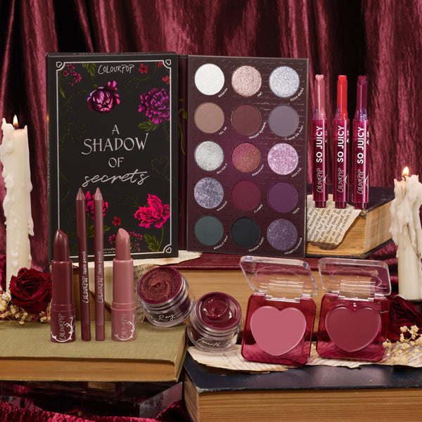 ColourPop Full Collection Set featuring A Shadow of Secrets Collection displayed on vintage books: 12‑pan plum‑toned shimmer eyeshadow palette, So Juicy glosses, lipsticks and liners, cream shadow pots, and heart‑shaped blushes amid candles and burgundy velvet.