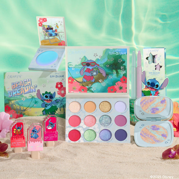Disney Lilo and Stitch Collection by ColourPop Full Collection Set displayed on sandy beach - 12-pan Beach Dreamin’ eyeshadow palette, lip gloss trio, two cream gel liners, marbled highlighter compact, and jelly highlighter in tropical Stitch packaging.