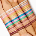ColourPop Socialite Shadow Stix swatched as a bold metallic teal eyeshadow stripe on three different skin tones, shown among other colorful Shadow Stix shades to highlight pigment payoff and smooth, creamy texture.