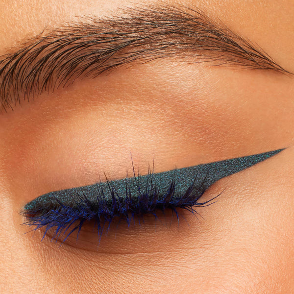 Winged eyeliner creates a sharp cat-eye on a closed eyelid, showcasing ColourPop Crème Gel Liner Out of the Blue - a metallic navy-blue shimmer - gliding smoothly along the lash line with vivid blue lashes in a close-up studio beauty shot.