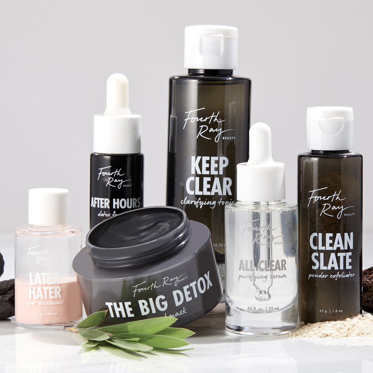 The Big Detox Mud Mask jar, lid off revealing charcoal-gray clay, sits centered among Fourth Ray Beauty skincare bottles on a white counter with green leaves; a ColourPop Face Mask for deep pore detox and clarifying.