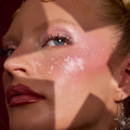 ColourPop Who-Hash glitter gel sparkling across the cheekbones and under-eye, scattering pink iridescent flecks and tiny white snowflakes over rosy blush and shimmer shadow; close-up holiday look with glossy berry lips and a snowflake earring.