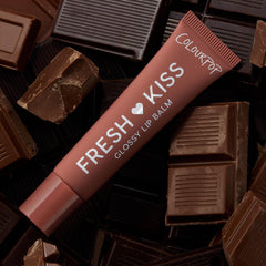 ColourPop Fresh Kiss glossy lip balm tube from the Get Rich ColourPop Glossy Lip Balm Trio lying diagonally on milk and dark chocolate bars, terracotta-brown packaging labeled Fresh Kiss Glossy Lip Balm and ColourPop in a chocolate-themed flat lay.