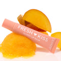 ColourPop Fresh Kiss glossy lip balm tube rests angled on a scoop of mango sorbet with mango slices, spotlighting the Pretty in Pastels ColourPop Glossy Lip Balm Trio; peach tube, hydrating shine on a clean white background.