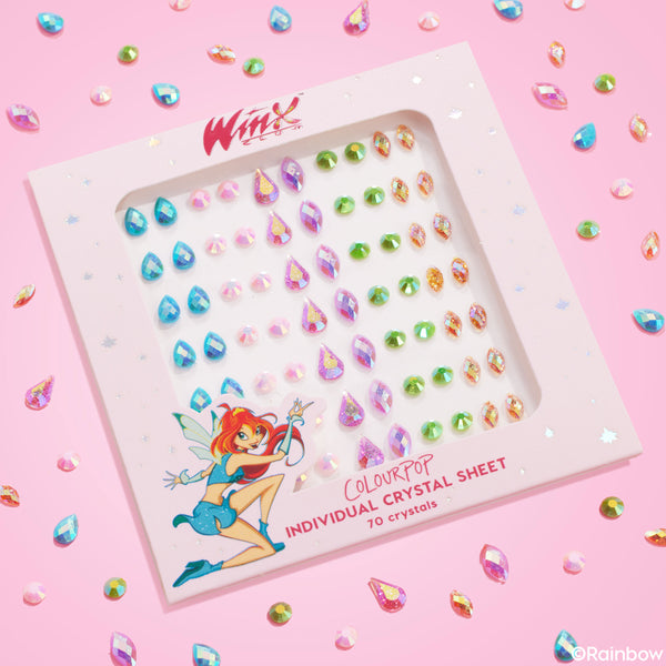 ColourPop Crystal Face Jewels Individual sheet displaying 70 self-adhesive face crystals - teardrop, round, and marquise - in iridescent pink, aqua, green, and gold, arranged in rows on Winx Club packaging, flatlay on a pink background with scattered gems.