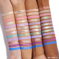 Swatched eyeshadows on three forearms display the ColourPop Prettiest in the Cosmos shadow palette set, 12 matte and shimmer shades from soft pinks, lilacs, and golds to vivid blue; labels show Twinkles, So Cosmic, Starlight, Aqua, Starry Sky.