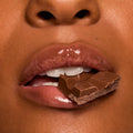 Glossy lips bite a piece of milk chocolate, showcasing rich shine in a chocolate-brown shade from Gloss Goals, a ColourPop Full Collection Set, in a tight beauty macro emphasizing a smooth, hydrated finish.