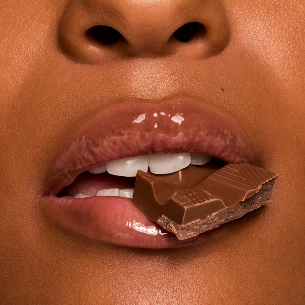 Glossy lips bite a piece of milk chocolate, showcasing rich shine in a chocolate-brown shade from Gloss Goals, a ColourPop Full Collection Set, in a tight beauty macro emphasizing a smooth, hydrated finish.