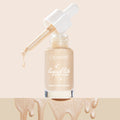 Sun O' Clock ColourPop Liquid Highlighter (Liquid Lite Glow Drops) in a frosted glass bottle, white dropper releasing a shimmering champagne drop, set against a soft beige backdrop with metallic glow drips along the bottom.