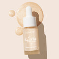 ColourPop Liquid Highlighter Sun O' Clock glow drops sit in a frosted glass dropper bottle with white cap, set atop swirling champagne-gold swatches that highlight its luminous shimmer on a soft beige backdrop.