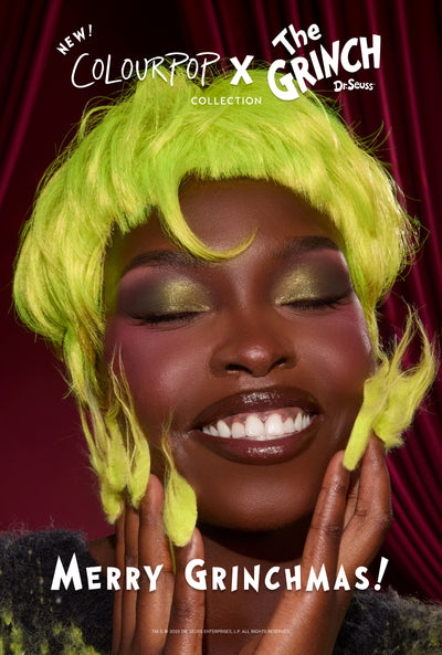 Model smiles with eyes closed, showing shimmery green eyeshadow and glossy lips, wearing a neon-green Grinch wig and fuzzy accents, promoting the ColourPop x The Grinch makeup collection against a red velvet backdrop with "Merry Grinchmas!" tagline.