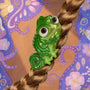 Tangled Pascal Hair Clip clasping a blonde braid, a shiny green chameleon-shaped barrette with large eyes from ColourPop Tools and Accessories, set against a whimsical purple floral painted backdrop.