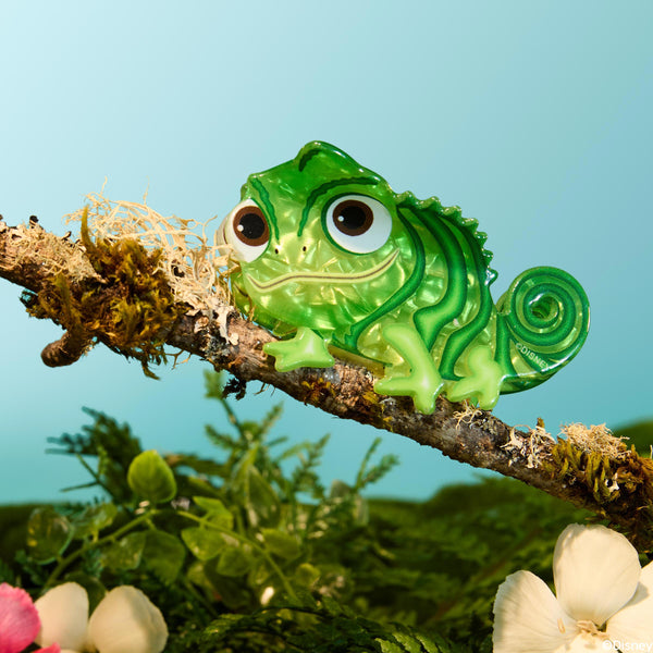 ColourPop Tangled Pascal Hair Clip perches on a mossy branch, a glossy green chameleon-shaped hair accessory with big brown eyes and a spiral tail in a stylized forest scene with flowers and blue sky - Disney Tools and Accessories.