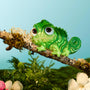 ColourPop Tangled Pascal Hair Clip perches on a mossy branch, a glossy green chameleon-shaped hair accessory with big brown eyes and a spiral tail in a stylized forest scene with flowers and blue sky - Disney Tools and Accessories.