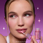 Model applies Gloss Me Up ultra glossy lip set in Heart to Get, holding the pink shimmery ColourPop-style tube against a purple starry background, showcasing high-shine nude-pink lips and matching soft glam makeup for a glossy finish.