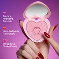 Hand with glossy red nails taps the shimmery pink Not Your Baby Super Shock Blush in a heart-shaped compact, highlighting Heart to Get bouncy pearlized formula, ultra-buildable blendability, and weightless glowy finish against a purple background.