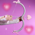 Hand draws a glittery silver bow with a pink heart-shaped arrow reading ColourPop, styled on a dreamy pink background to tease the Not Your Baby Heart to Get Super Shock Blush Valentine-inspired makeup collection.