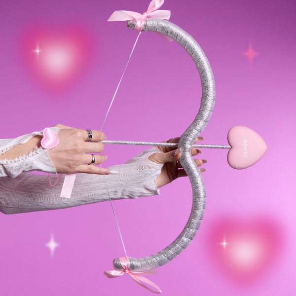 Hand draws a glittery silver bow with a pink heart-shaped arrow reading ColourPop, styled on a dreamy pink background to tease the Not Your Baby Heart to Get Super Shock Blush Valentine-inspired makeup collection.