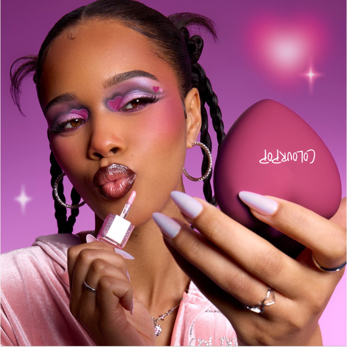 Model applies shimmery pink gloss while holding a heart-shaped ColourPop compact, showing bold purple and pink heart eye makeup that highlights the Heart to Get Full Collection full collection set against a dreamy pink background.