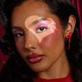 Model wears shimmering pink lids and icy inner-corner highlight created with the ColourPop Liked Christmas a Lot… Shadow Stix Set, accented by blush, glossy red lips, a heart-shaped light, and a red bow earring against a deep red backdrop.