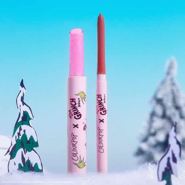 Colourpop Pinky Who So Juicy Plumping Gloss Stick stands uncapped beside a coordinating lip liner, revealing a glittery pink bullet; both in The Grinch x ColourPop pink tubes, set in a snowy pastel-blue holiday scene with cartoon trees.