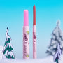 Colourpop Pinky Who So Juicy Plumping Gloss Stick stands uncapped beside a coordinating lip liner, revealing a glittery pink bullet; both in The Grinch x ColourPop pink tubes, set in a snowy pastel-blue holiday scene with cartoon trees.