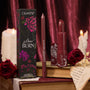 ColourPop Lux Lipstick in Slow Burn, a deep rose‑burgundy bullet with matching lippie pencil, stands beside a floral box on stacked books, framed by red roses and melting candles against a rich burgundy velvet backdrop.