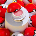 Type Advantage ColourPop Lip Set lip mask opens to reveal shimmery pink balm in a Poké Ball–inspired jar, displayed on a white pedestal and encircled by glossy red-and-white spheres.