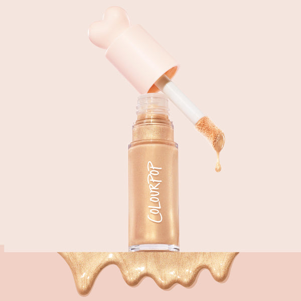 ColourPop Liquid Highlighter in Immediately Yes drips from a doe‑foot applicator above a clear tube, revealing a shimmery champagne‑gold formula; a pale pink heart‑shaped cap and glossy gold spill sit on a soft blush background.