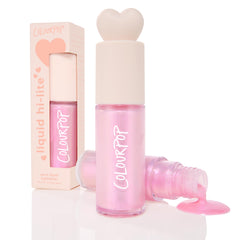ColourPop Liquid Highlighter in Pink Pony stands upright with a heart-shaped cap as a second bottle spills shimmery iridescent pink onto a white surface, with the peach outer box behind; dewy finish, 10 g/0.35 oz.