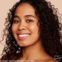 Model smiling in a close-up, wearing ColourPop Pretty Fresh Tinted Moisturizer in Medium Dark 15 W, showing dewy, even coverage and natural finish against a soft beige background.
