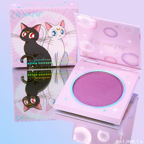 Moon Kingdom ColourPop Pressed Powder Highlighter compact displays a shimmering lavender pan in pastel pink, star-accented packaging, beside the matching box featuring Sailor Moon’s cats Luna and Artemis on a reflective surface.