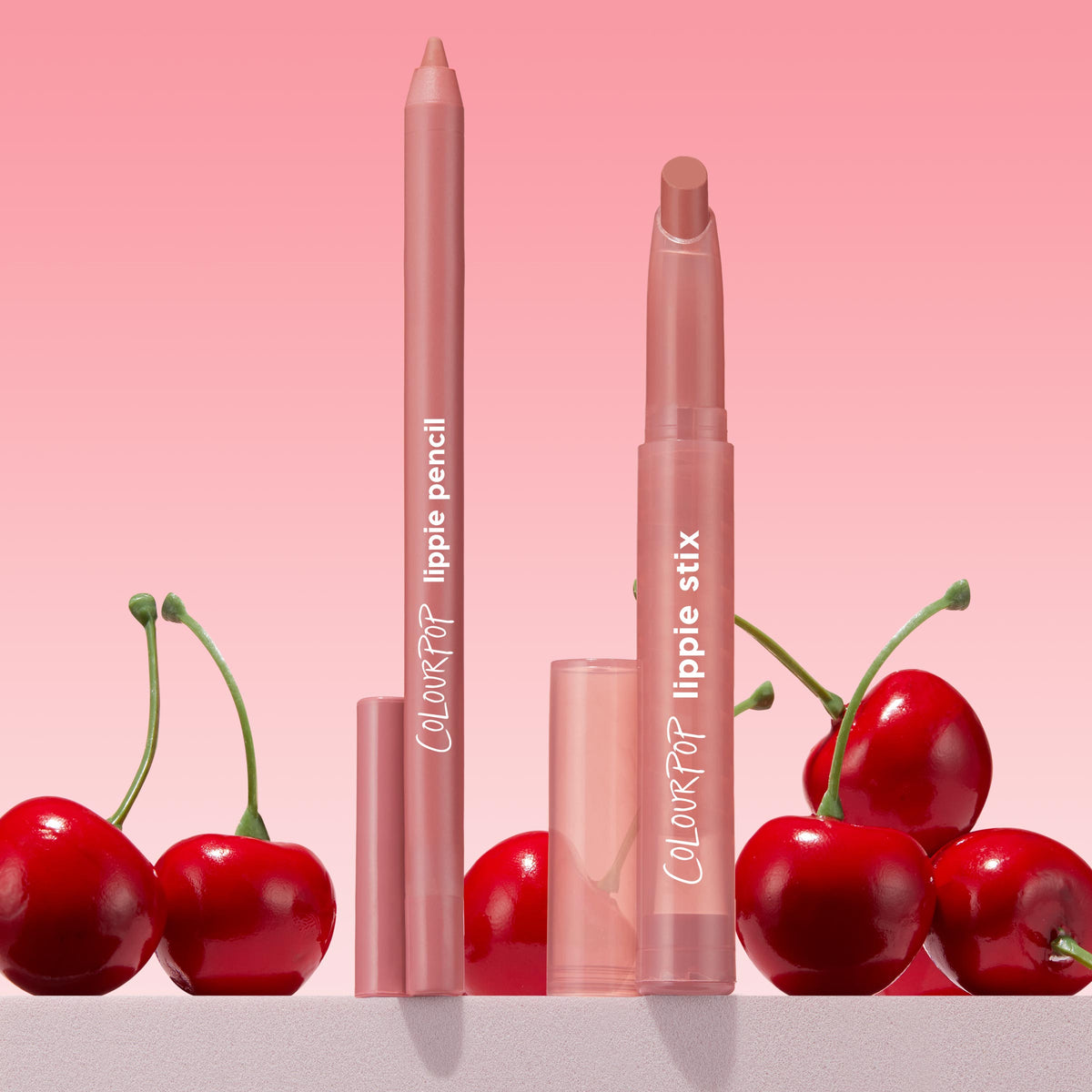 ColourPop Lippie Pencil "Oh Snap" stands uncapped with a creamy soft pink-nude tip, paired with matching Lippie Stix, on a countertop with glossy red cherries against a pink gradient backdrop - ideal for precise, long-wear lip lining.