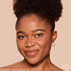 Model wearing ColourPop Pretty Fresh Tinted Foundation Balm in Dark 18, smiling in a studio close-up that showcases smooth, even, dewy skin with natural coverage, small hoop earring visible, against a beige backdrop for accurate shade reference.