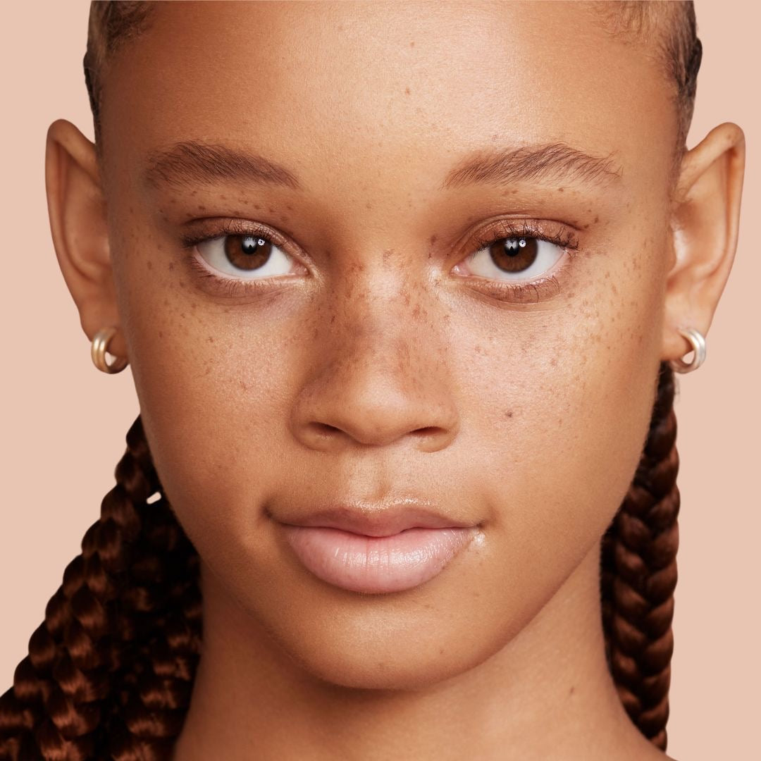 ColourPop Pressed Powder Bronzer in Silver Strand Beach warming a model’s complexion, showing a soft sun-kissed glow on the cheeks in a close-up studio portrait with freckles, glossy lips, braided hair, and hoop earrings against a peach background.