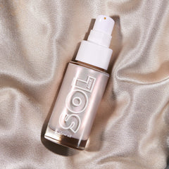 Moonstone SOL Glow Oil bottle tilts on champagne silk, its frosted glass and white pump catching light with a pearlescent glow; ColourPop SOL Glow Oil for luminous body shine in a sleek pump design.