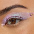 Individual ColourPop Crystal Face Jewels accent a close-up eye, with an iridescent teardrop gem at the outer wing and a small round pink opal gem below the lower lash line, complementing shimmering lavender winged eyeshadow and natural lashes.