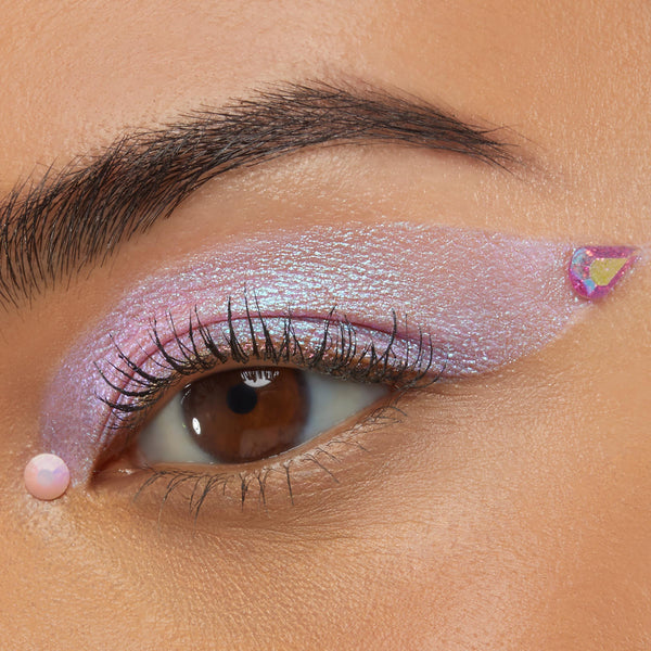 Individual ColourPop Crystal Face Jewels accent a close-up eye, with an iridescent teardrop gem at the outer wing and a small round pink opal gem below the lower lash line, complementing shimmering lavender winged eyeshadow and natural lashes.