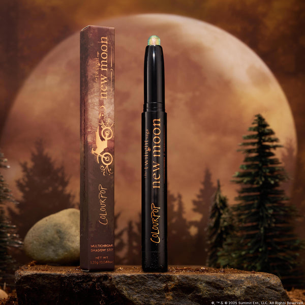 ColourPop Shadow Stix 'It’s Always Been Him' stands uncapped, revealing a shimmery green multichrome tip on a black twist-up stick with gold lettering, beside its box on a rock, framed by pine trees and a glowing full moon.