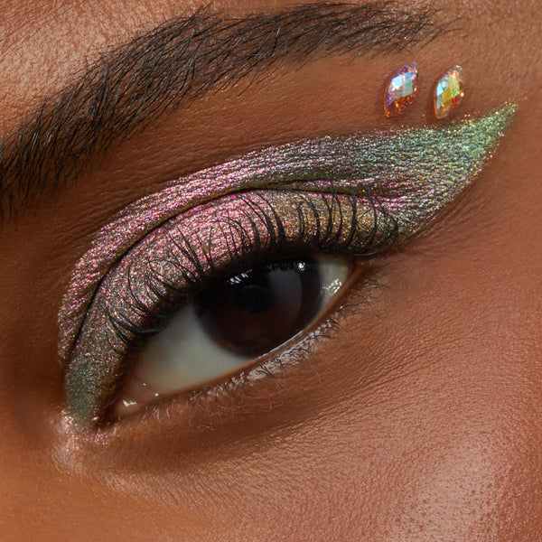 Two iridescent teardrop gems adhere above the outer eye, showcasing the Individual ColourPop Crystal Face Jewels with winged multichrome pink‑green shimmer eyeshadow and long lashes for festival‑ready makeup.