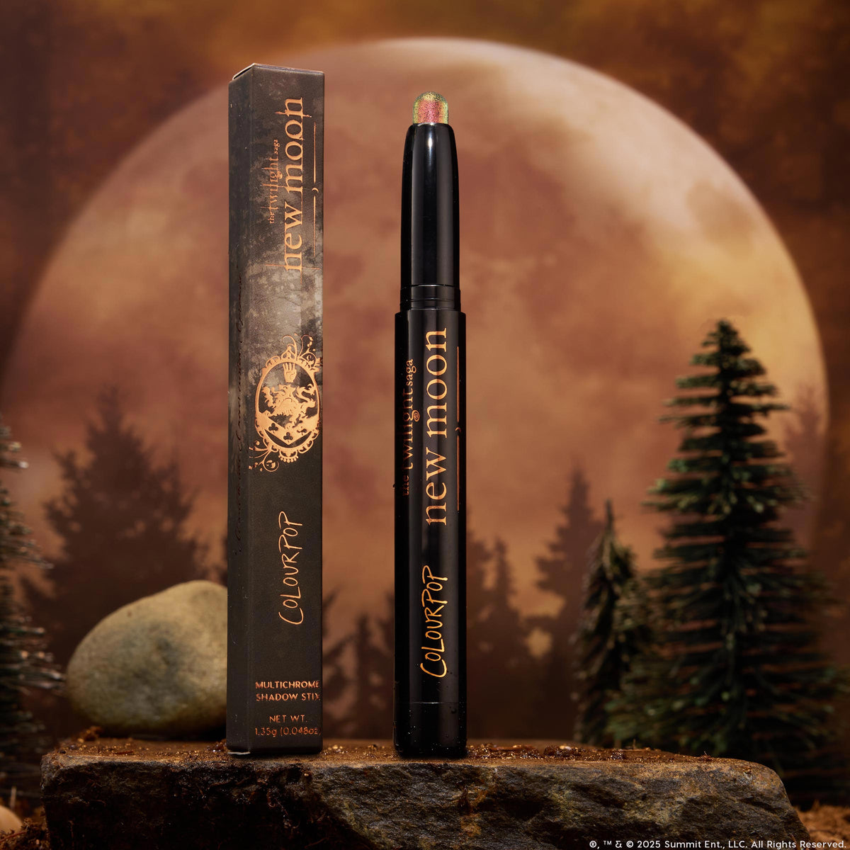 Vampire Girl ColourPop Shadow Stix stands uncapped, showing a pink‑green multichrome tip on a sleek black tube with gold lettering, its box beside it, set on a rocky surface with pine trees and a glowing full moon background.